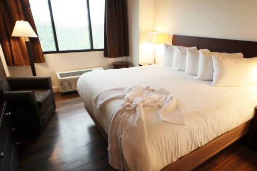 Cozy hotel room featuring a large bed, armchair, and window with curtains, illuminated by a bedside lamp.