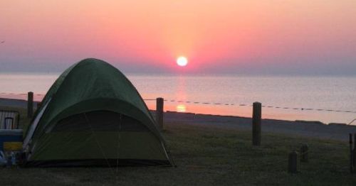 A green tent sits on grass by the shore, with a vibrant sunset reflecting on calm waters in the background.