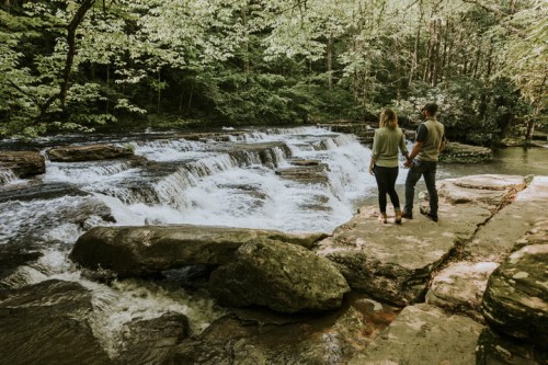 A couple stands by a flowing river with waterfalls, surrounded by lush green trees in a serene natural setting.