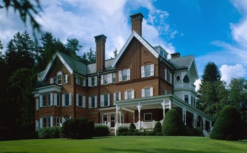 A large, elegant brick mansion with a porch, surrounded by lush greenery and trees under a blue sky.