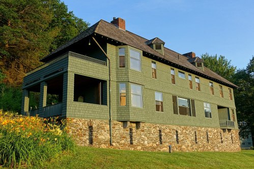 A large green house with multiple windows and a stone foundation, surrounded by greenery and flowers in the foreground.