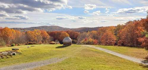 A scenic view of rolling hills and vibrant autumn foliage under a partly cloudy sky. A path leads to a dome structure.