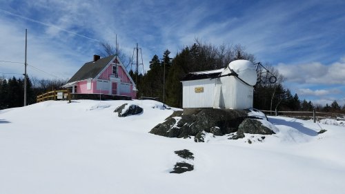 A pink house and a white structure sit on snowy ground, surrounded by trees under a blue sky with clouds.