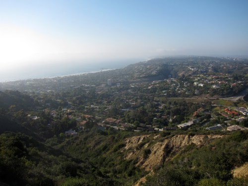 A panoramic view of a coastal landscape with hills, homes, and the ocean in the distance under a clear sky.