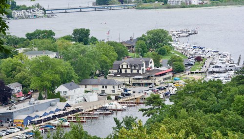 Aerial view of a marina with boats, green trees, and buildings along the water's edge.