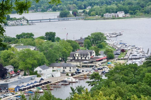 Aerial view of a marina with boats, green trees, and a river, featuring a bridge in the background.