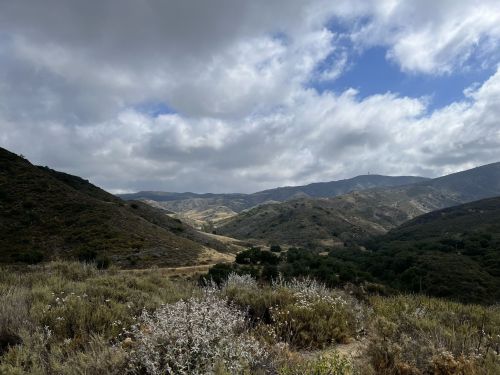 A scenic view of rolling hills and valleys under a partly cloudy sky, with patches of greenery and dry grass.