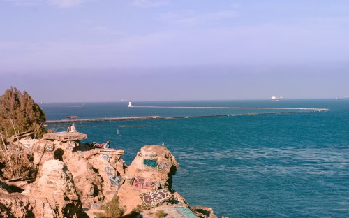 A rocky coastline with colorful graffiti, overlooking a calm blue sea and distant boats on the horizon.