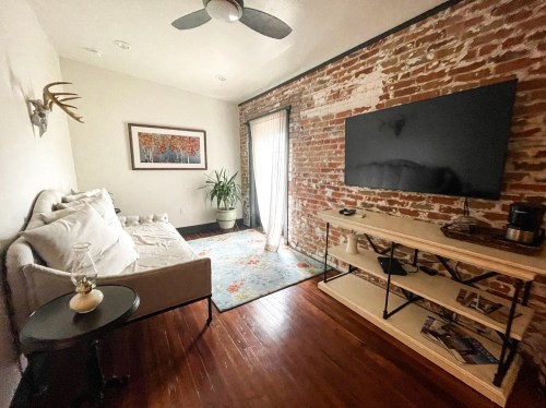 Cozy living room with exposed brick walls, a sofa, TV, and a plant, featuring a colorful rug and natural light.