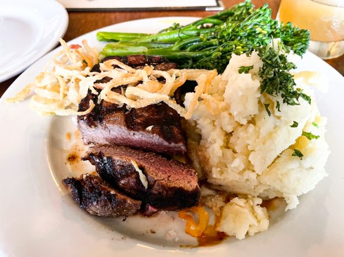 A plate featuring grilled steak, creamy mashed potatoes, sautéed greens, and crispy onion strings.