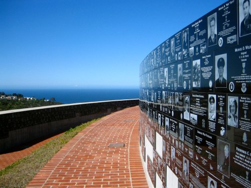 A curved memorial wall with plaques honoring individuals, overlooking a blue ocean under a clear sky.