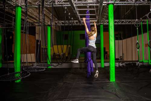 A person climbs a purple fabric in a gym with green pillars and various training equipment.