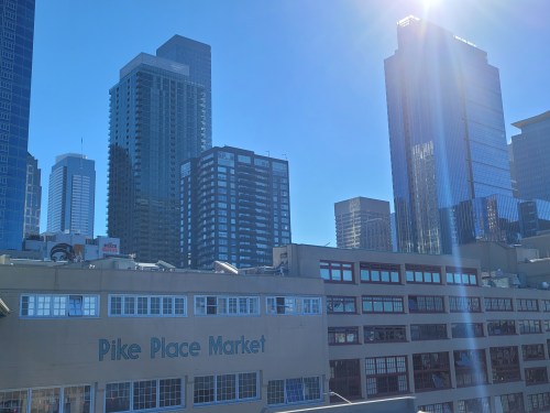 View of Pike Place Market with modern skyscrapers and a clear blue sky in the background. Sunlight reflects off buildings.