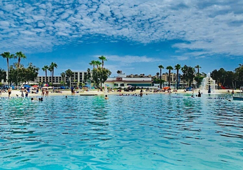 A sunny pool scene with people swimming and lounging, surrounded by palm trees and a blue sky with clouds.