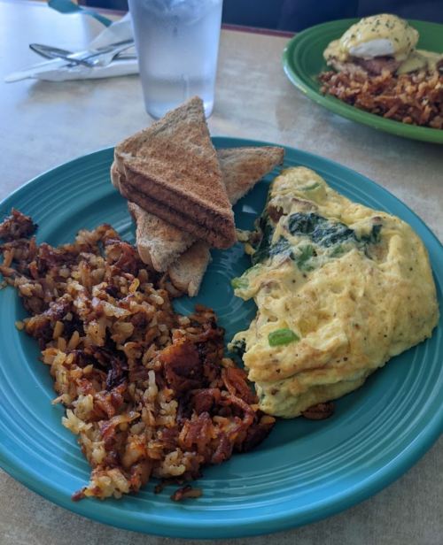 A blue plate with a spinach omelette, hash browns, and toast, alongside a glass of water in a casual dining setting.