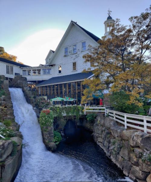 A picturesque building beside a waterfall, surrounded by trees and outdoor seating, under a clear blue sky.
