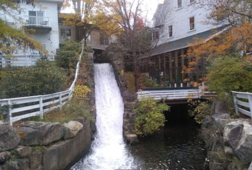 A waterfall flows into a serene pond, surrounded by autumn foliage and quaint buildings.