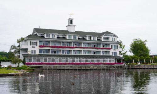 A charming white hotel with flower-filled balconies, overlooking a calm lake with ducks swimming nearby.