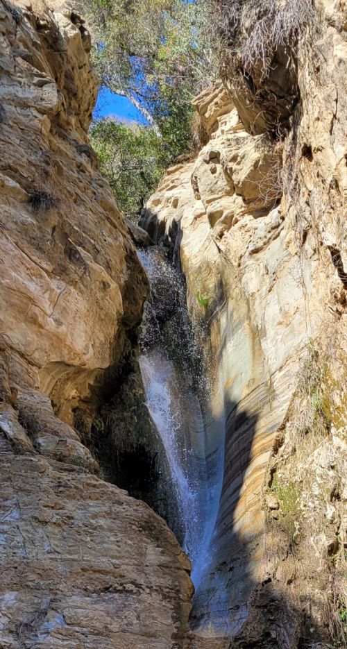 A waterfall cascading down rocky cliffs, surrounded by greenery and a clear blue sky above.
