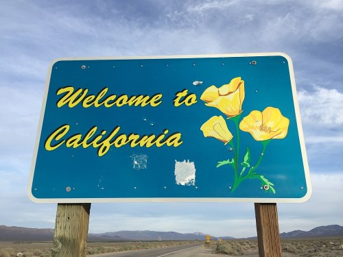 A blue welcome sign for California featuring yellow flowers and a cloudy sky in the background.