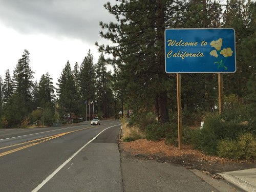 A roadside sign reading "Welcome to California" surrounded by tall pine trees and a cloudy sky.