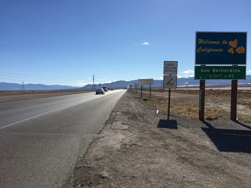 A highway scene with a "Welcome to California" sign and mountains in the background under a clear blue sky.