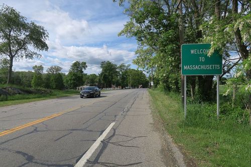 A road sign reading "Welcome to Massachusetts" alongside a tree-lined street with a car driving by.