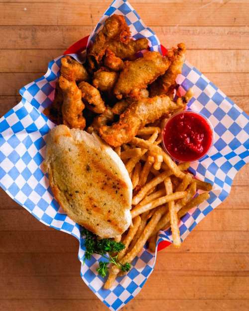 A basket of fried fish, golden fries, a toasted bun, and a side of dipping sauce on a checkered paper.