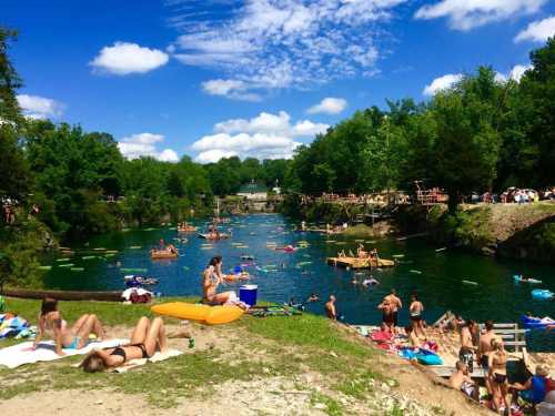 A sunny day at a river with people lounging on the shore and floating on rafts, surrounded by lush green trees.