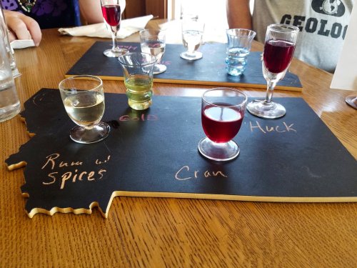 A wooden board shaped like a state with various colorful shot glasses labeled with names like "Cran" and "Huck."