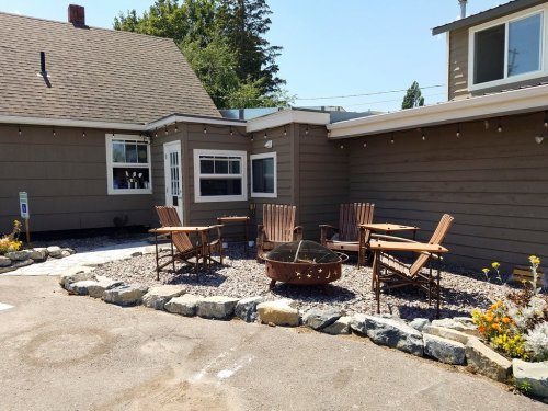 Outdoor seating area with wooden chairs and a fire pit, surrounded by gravel and flowers. Sunny day.