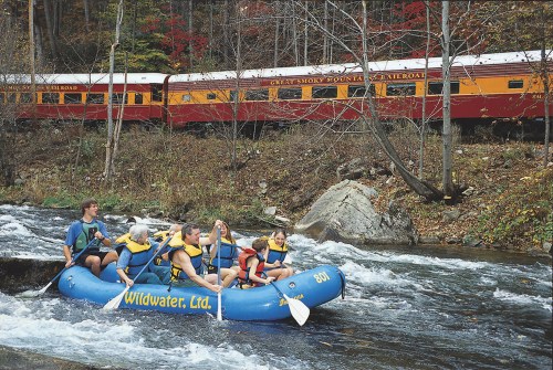 A group of people in a blue raft navigating a river, with a colorful train passing by in the background.