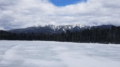 Snow-capped mountains rise above a frozen lake, surrounded by evergreen trees under a cloudy sky.