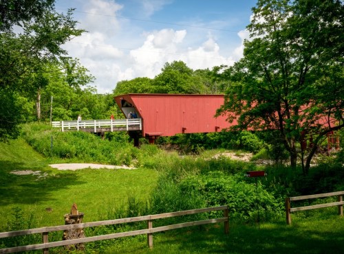 A red covered bridge surrounded by lush greenery and trees, with people walking on a nearby path.
