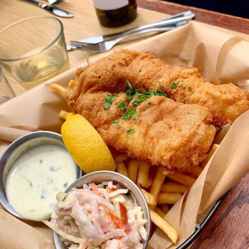 A basket of golden fried fish fillets, fries, coleslaw, and a lemon wedge, garnished with parsley.