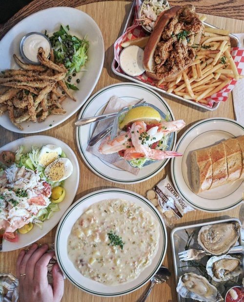 A table spread with various seafood dishes, including shrimp cocktail, fried oysters, a sandwich, and clam chowder.