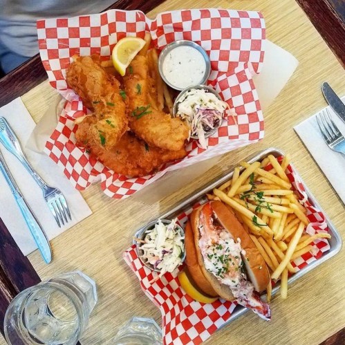 A table with two baskets of food: fried fish with lemon, coleslaw, and a lobster roll with fries.