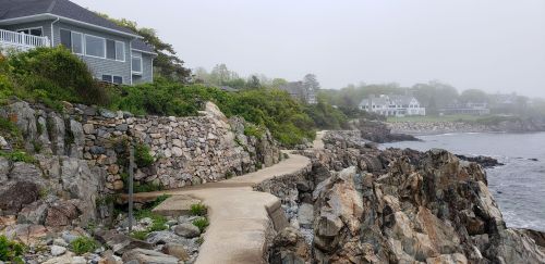 A foggy coastal scene featuring a rocky path along the shore, with houses visible in the background.