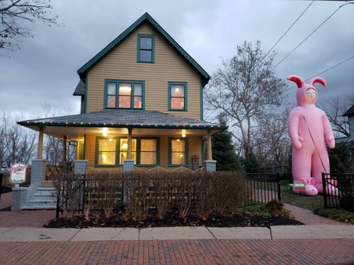 A large pink inflatable bunny stands next to a charming yellow house with festive lights and a cloudy sky.