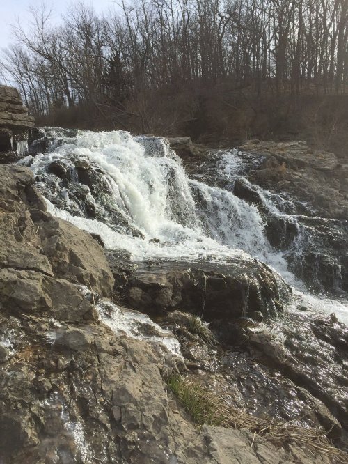 A cascading waterfall flows over rocky terrain, surrounded by bare trees and a serene natural landscape.