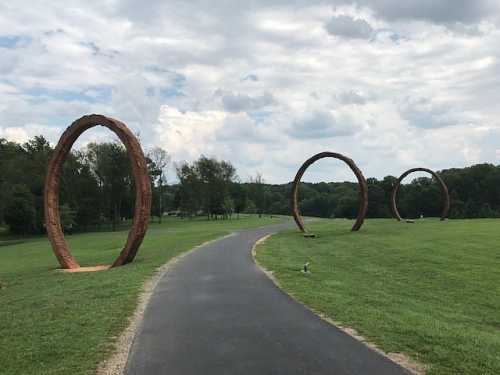 Three large wooden rings stand along a grassy path under a cloudy sky, surrounded by trees.