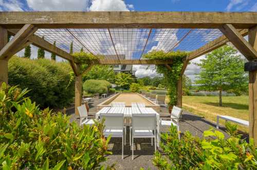 A serene outdoor space with a wooden pergola, white furniture, and lush greenery under a blue sky with fluffy clouds.