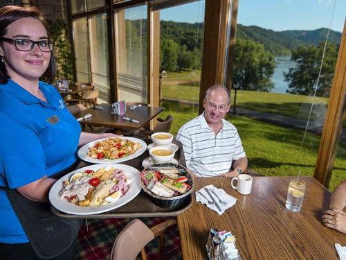 A waitress serves two trays of food to a smiling man at a table with a scenic view of a lake and mountains.