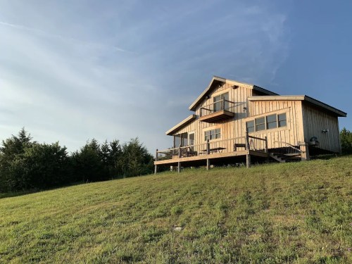 A wooden house on a grassy hill, surrounded by trees, under a clear blue sky.