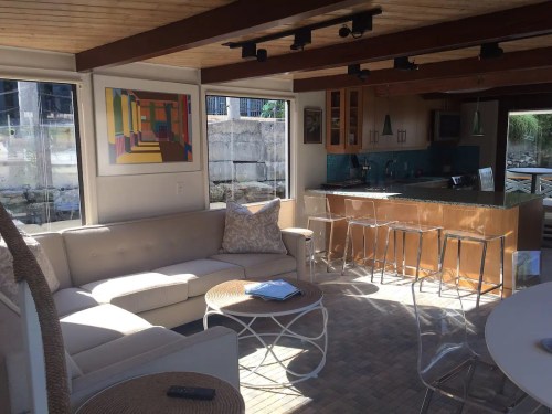 Bright, modern living space with a sofa, coffee table, and kitchen area featuring large windows and colorful artwork.