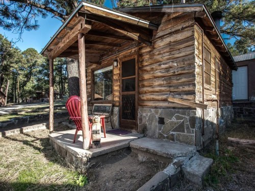 A rustic wooden cabin with a stone foundation, featuring a porch and a red chair in a forested area.