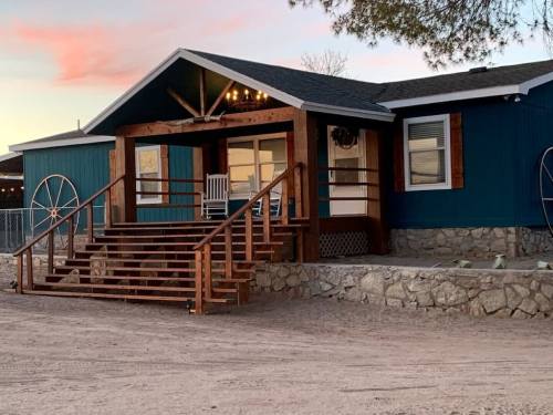 A blue house with a wooden porch and steps, featuring a rocking chair and stone foundation, set against a sunset sky.