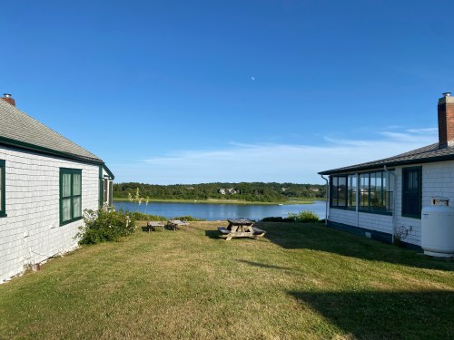 A serene view of a grassy area between two houses, overlooking a calm lake under a clear blue sky.