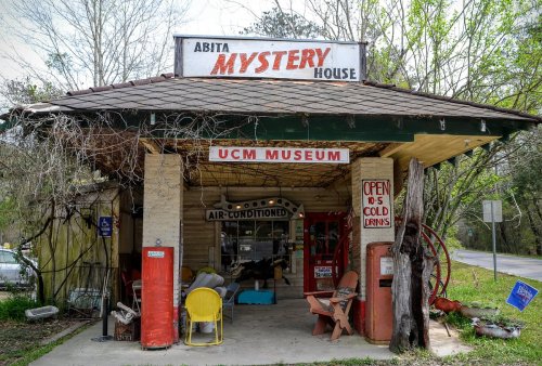A quaint roadside building with a sign reading "Abita Mystery House" and "UCM Museum," featuring vintage decor and seating.