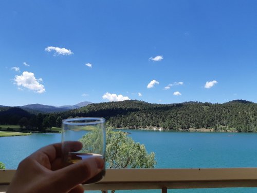 A hand holding a glass overlooks a serene lake surrounded by green hills and a clear blue sky with fluffy clouds.